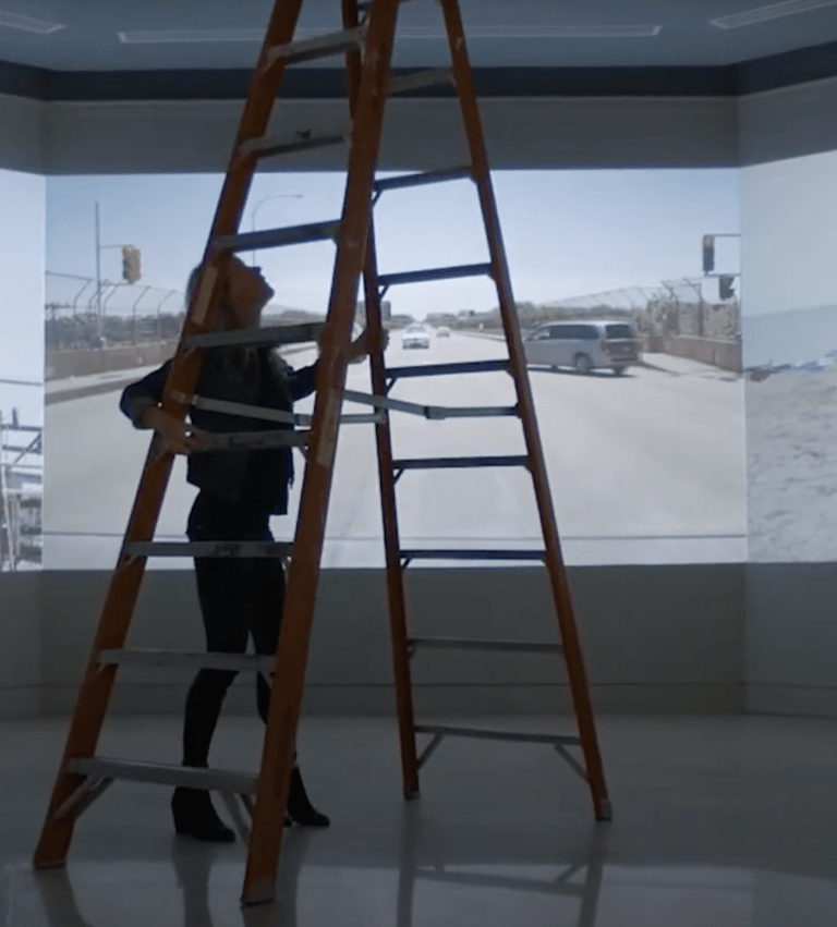 A person moves a tall ladder through a gallery space with large photographs on the walls.