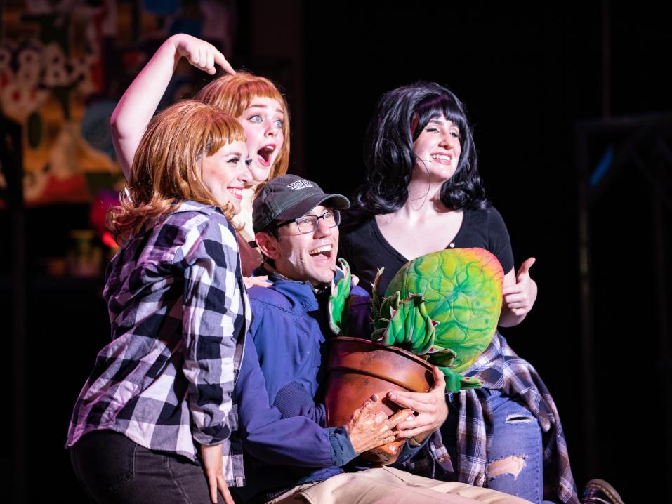 Four students appear on stage in a performance of Little Shop of Horrors. The actor playing Seymour holds a prop venus fly trap as they sing.