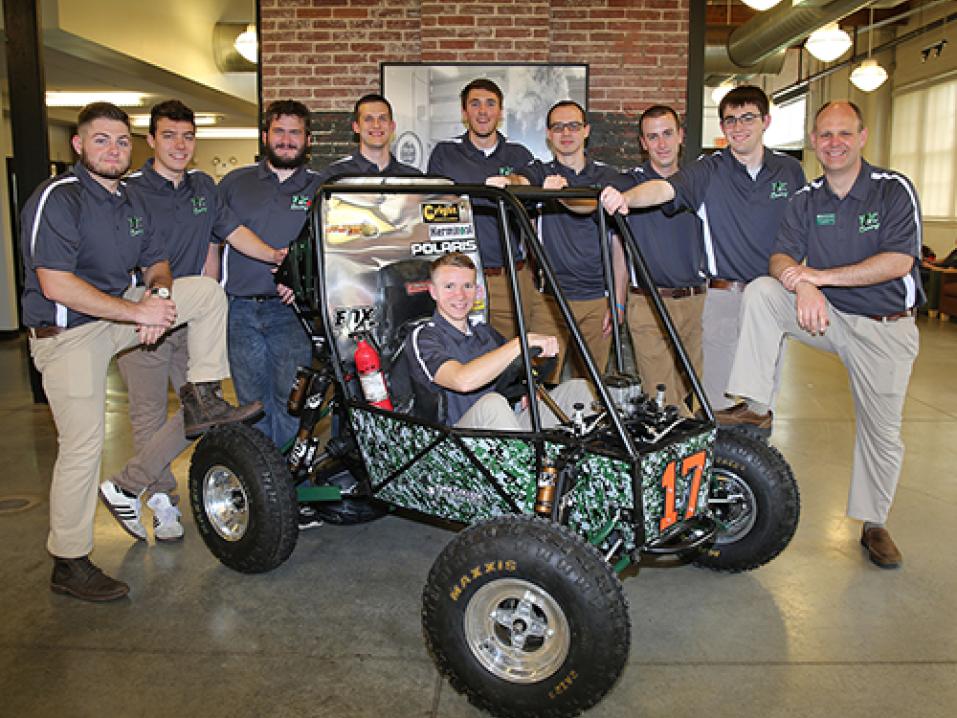 A group of student racers stand around their Baja SAE racecar. One student sits in the driver's seat.