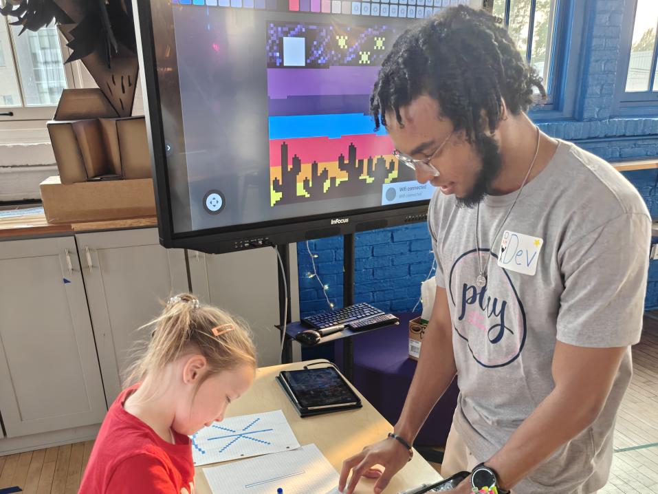 A young girl and an instructor collaborate on a pixel art activity using graph paper at a colorful, interactive learning station with a digital display in the background.