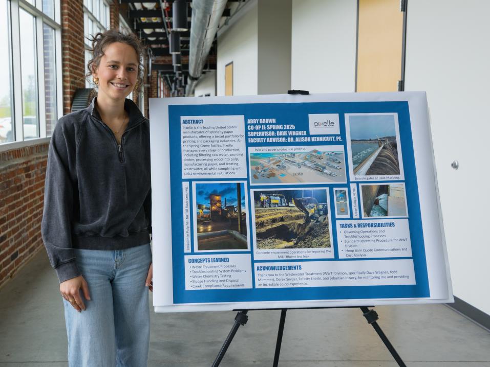 Abby Brown stands next to her co-op poster in a hallway with a brick wall and windows behind her.