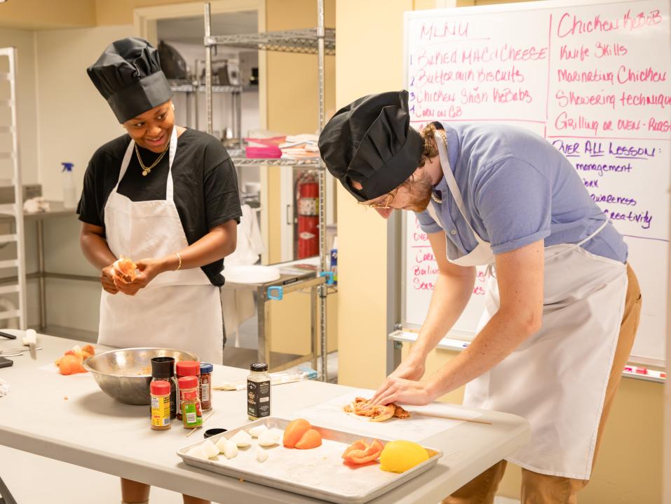 Two people prepare food in a kitchen wearing aprons and hats. A large whiteboard with writing is behind them.