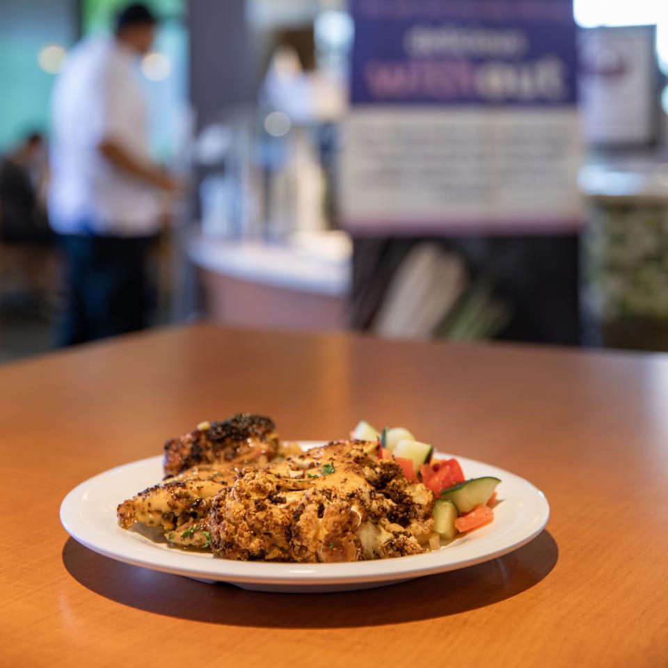 A plate of chicken and vegetable side sits on a dining hall table in front of the Delicious Without Station.