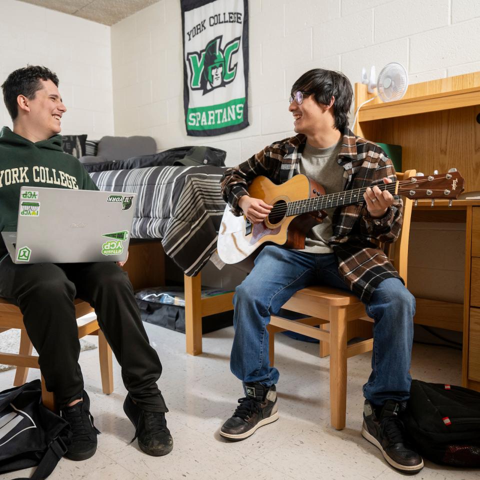 Two students sit in a residence hall dorm room, playing guitar and working on a laptop.