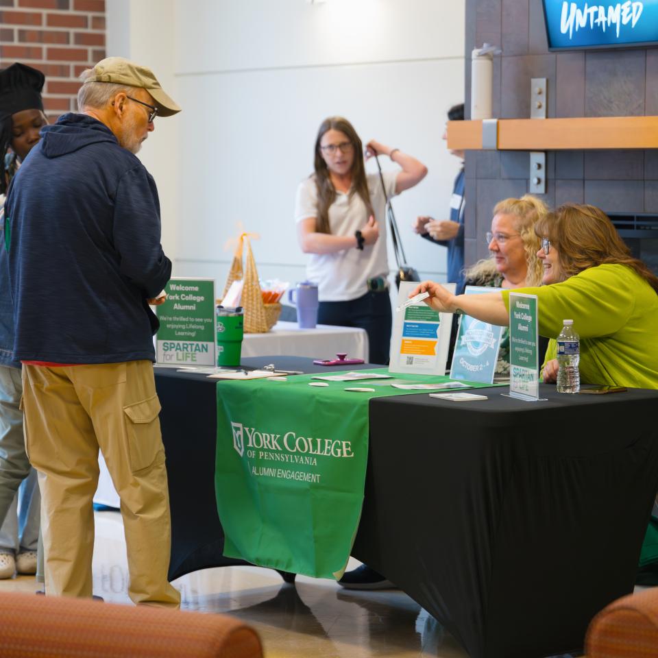 Two women sit at a table that reads, "York College Alumni Engagement" and hand out information to a man in front of them.