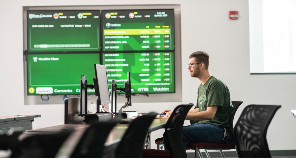 A student sits in a classroom with large computer monitors in front of him on the desk and huge 4-monitor screen on the wall behind him, displaying market data.