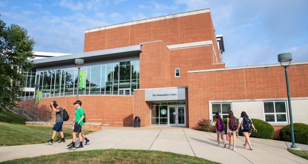 An exterior view of the Humanities Center on a sunny, blue-sky day. Students walk on the sidewalk in front of the building.