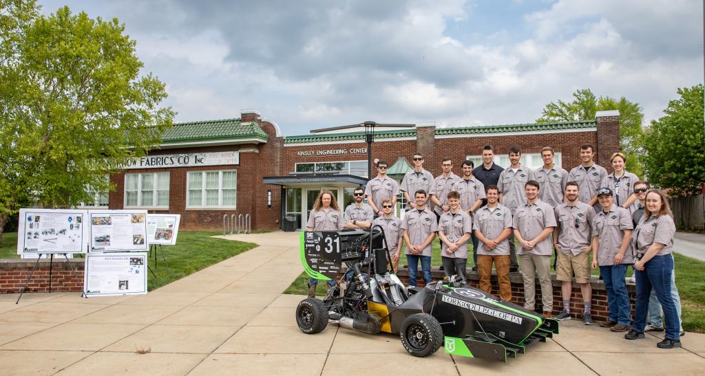 A group of students wearing matching uniforms stand behind their racecar in front of the Kinsley Engineering Center.