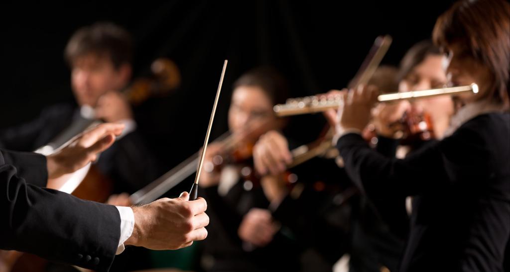 A conducter's hand is visible in front of an orchestra mid-performance.
