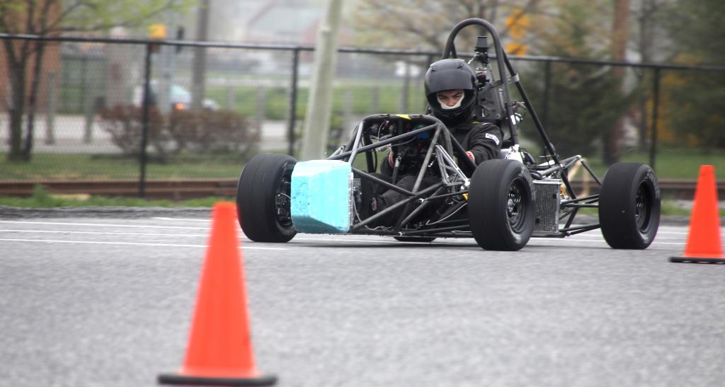 A student in a helmet and protective gear drives a formula SAE racecar through traffic cones in a parking lot.