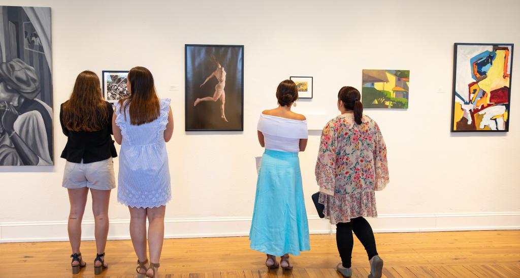Four people stand in front of a gallery wall where paintings are hung.