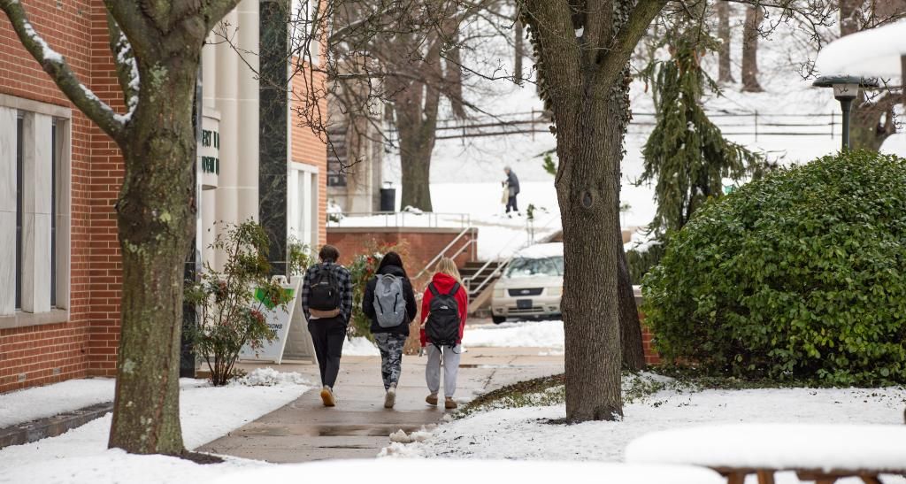 Three students walk across a campus sidewalk on a snowy day.