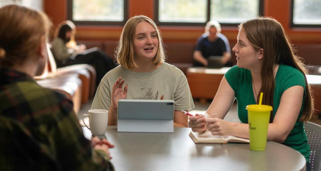 Three students sit around a table in a common area, chatting as they drink coffee and glance at an iPad.