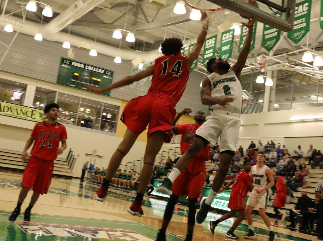 A York College men's basketball player jumps to throw a layup while a member of the opposing team jumps to try to block the shot.