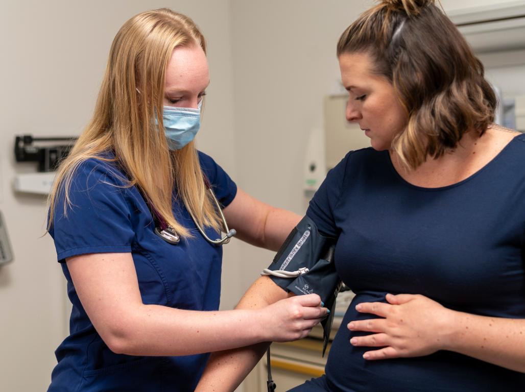 A nursing student wearing a medical mask takes the blood pressure of a pregnant woman in a clinical setting.