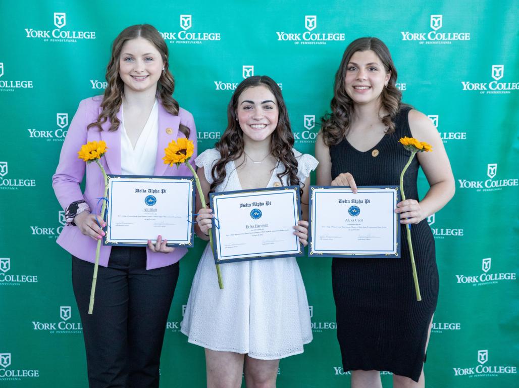 Three students smile in front of a York College logo backdrop, holding certificates and sunflowers.