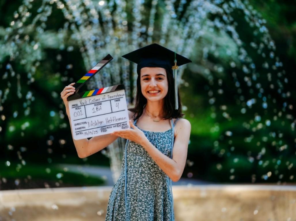 Ashley Hettleman poses in front of the campus fountain wearing her graduation cap and holding an open clapboard. 