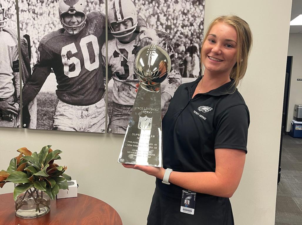 Ashley Hudak smiles while holding up a Vince Lombardi trophy in an office with a large football photo on the wall.