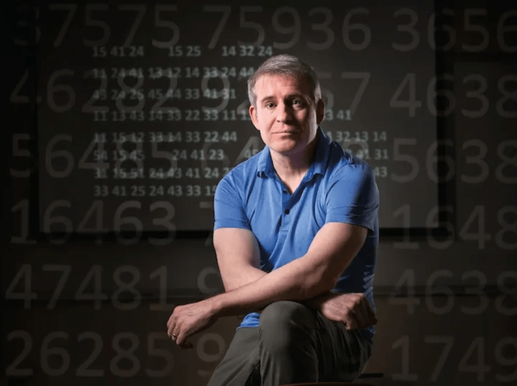 Craig Bauer poses for a headshot in front of a black backdrop covered in white chalk-style written formulas.