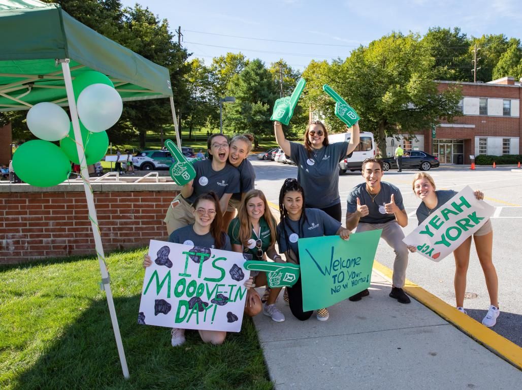 Students smile and wave as they stand by a tent holding signs with welcome messages.