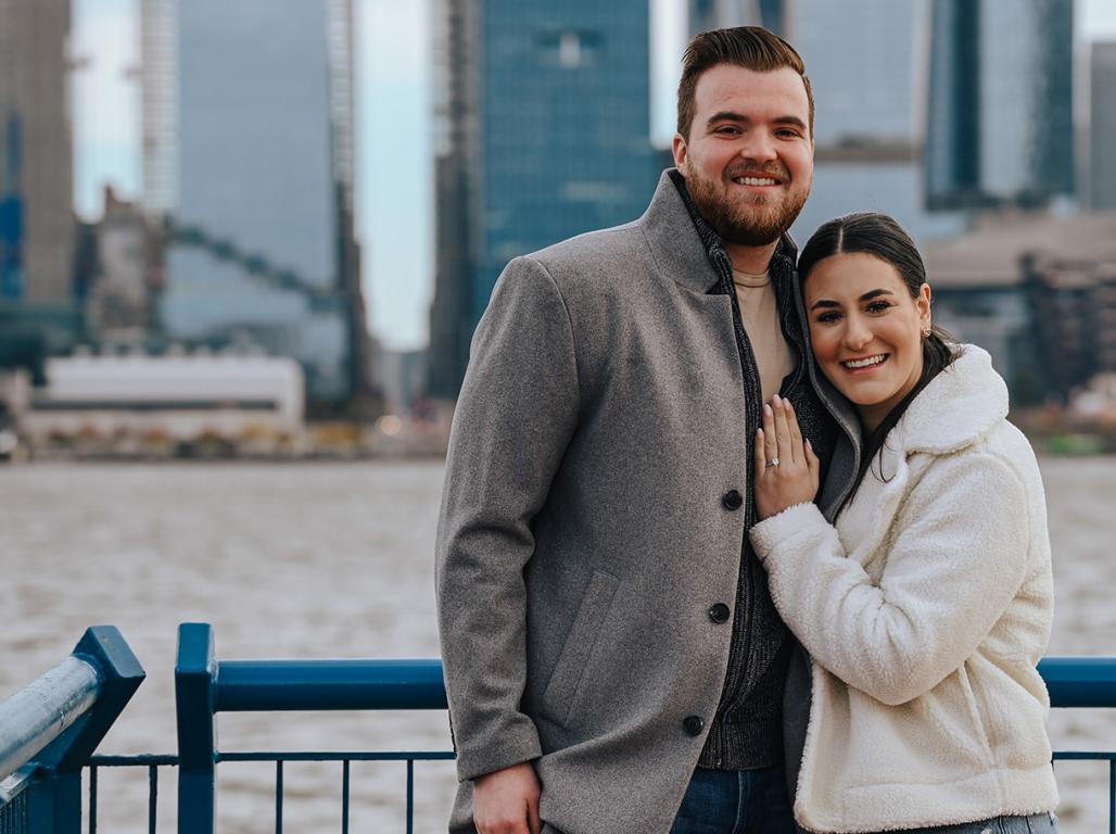 Gabriella Citakian and Tom Sweezy class of '17 pose smiling in front of a body of water separating them from a cityscape. Gabriella shows her engagement ring on her left hand. 