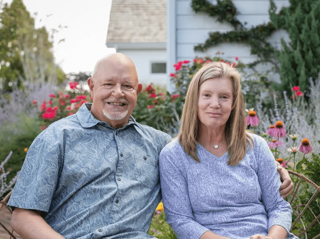 George Glatfelter II and Beverly Glatfelter sit for a photo in a garden. Red flowers and greenery are visible behind them.