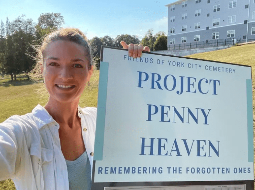 Jamie Noerpel smiles as she stands in a field, holding a sign that reads "Friends of the York City Cemetery - Project Penny Heaven - Remembering the Forgotten Ones."