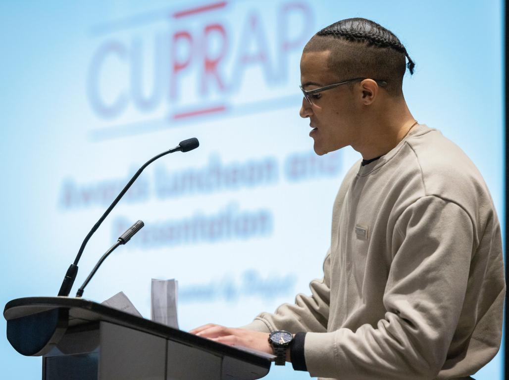 Kai O'Brien speaks at a podium on stage in front of a backdrop with the CUPRAP logo.