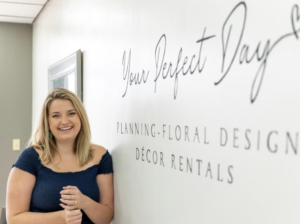 Leah Barnes, owner of Your Perfect Day LLC, stands smiling next to her hand painted business sign on a wall inside her store.