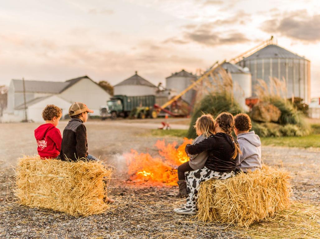Children on a farm sit on bales of hay around a fire. 