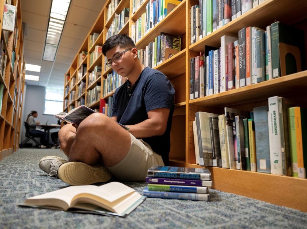 A student sits on the floor, back to a library stack, reading. Several open books are laid out around him.