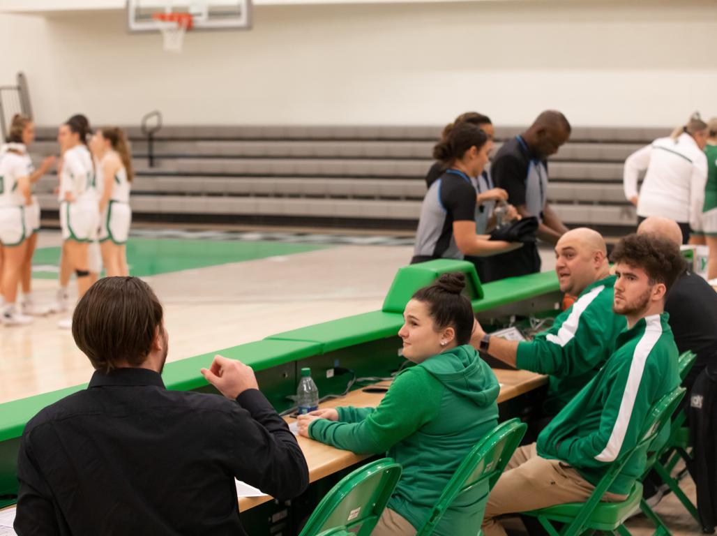 Sports officials converse at a table alongside the court during a Women's Basketball game.