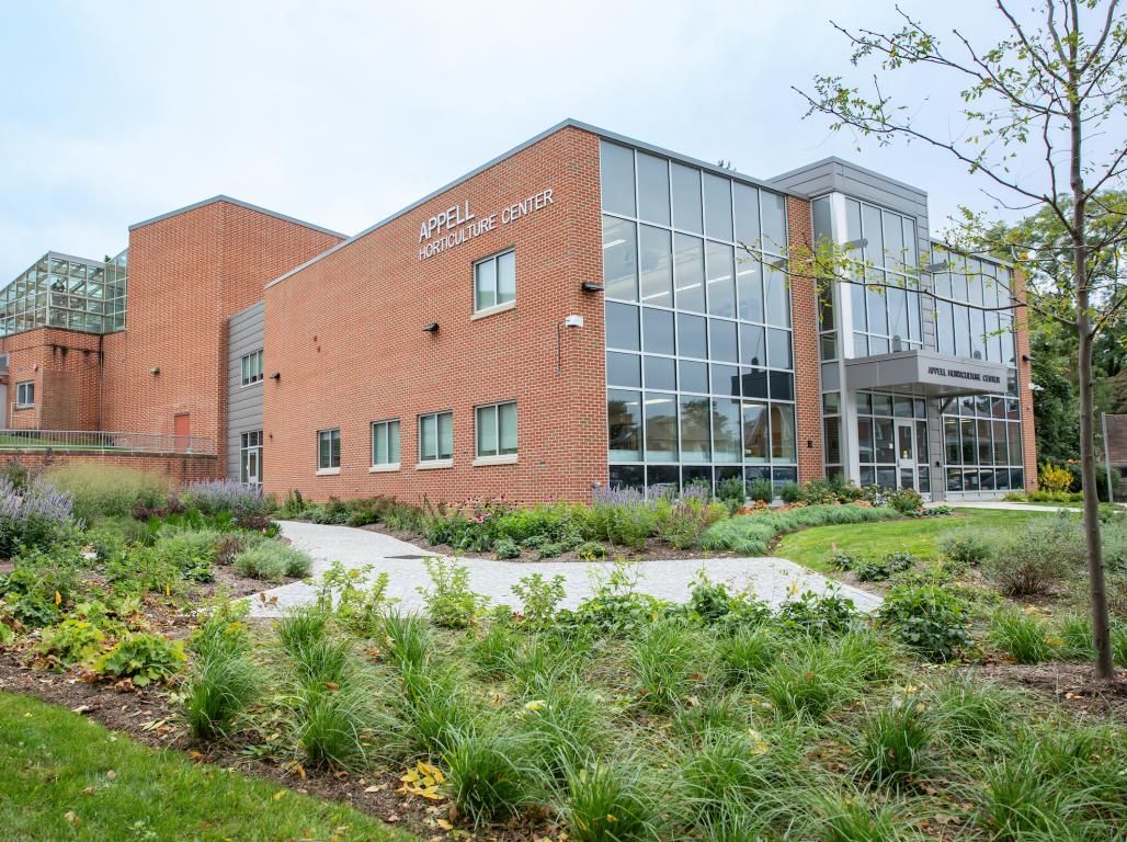 An exterior view of the Appell Horticulture Center shows landscaping with dozens of plans in front of a brick and glass-front building.