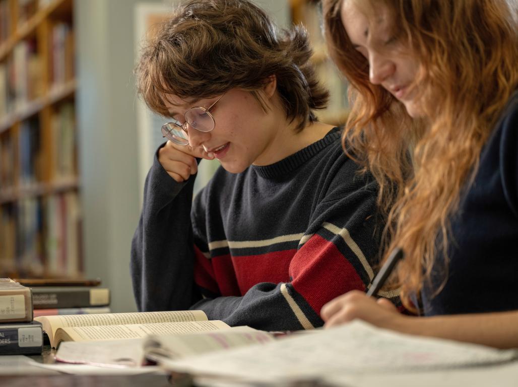 Two students study side by side as they look through papers on a desk in the library.