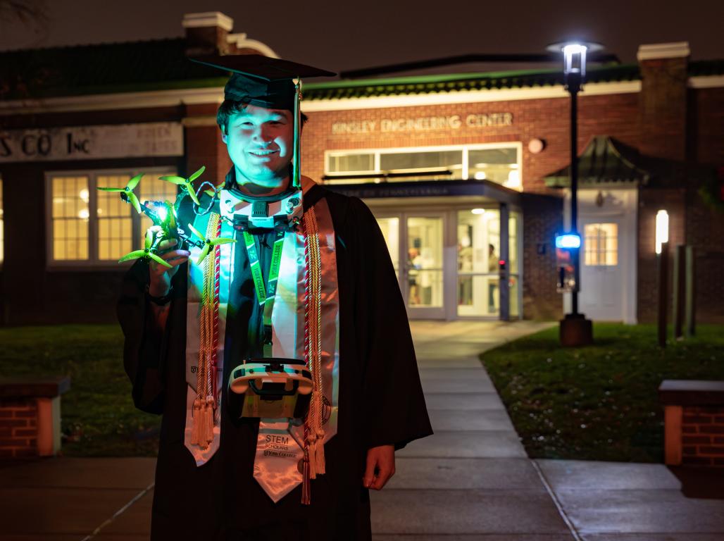 December graduate, Alex Suarez, stands in his cap and gown with a drone hovering in the dark.