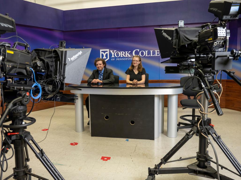 Two Mass Communication students sitting at the news desk with film cameras around them.