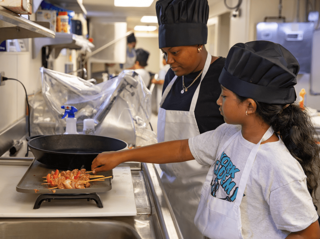 A student cooks a meal at the CCE while a teacher looks on.