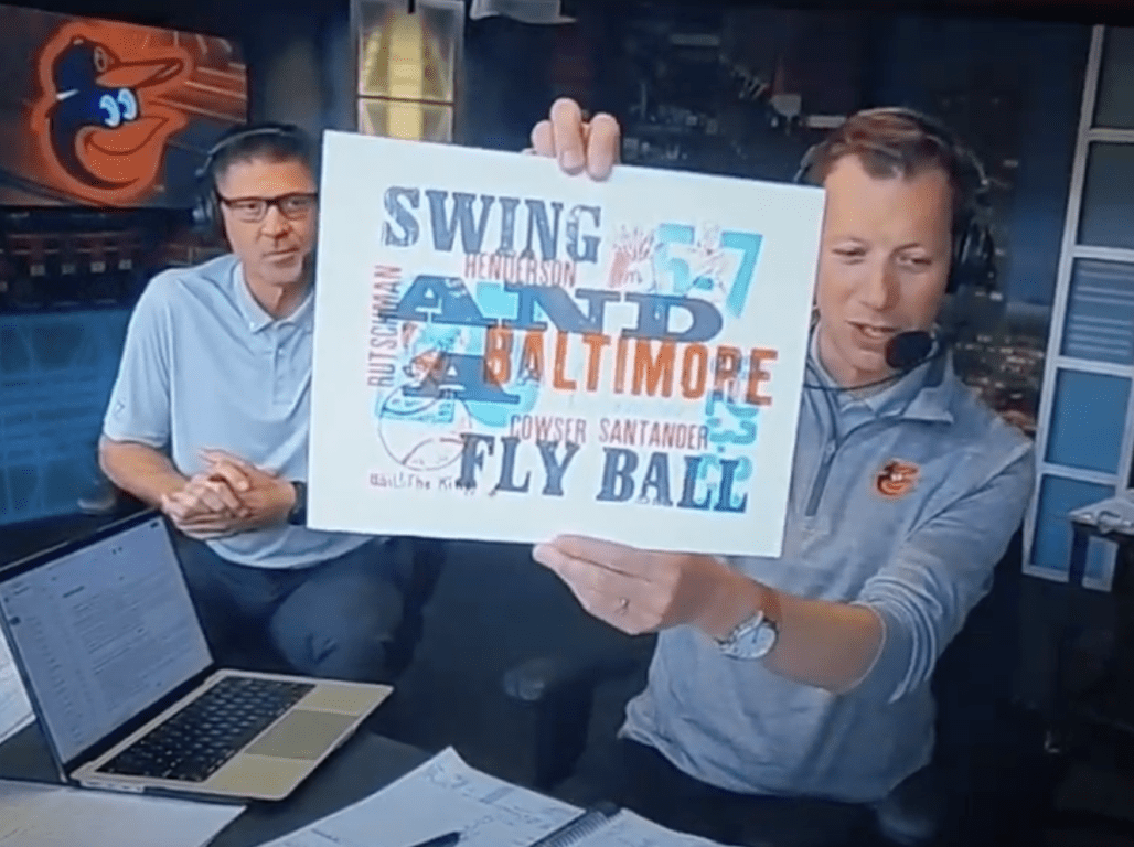 A sports broadcaster holds a colorful print up for the camera during a broadcast.
