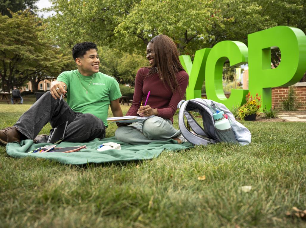 Two students at York College smile and study on a green blanket in front of large YCP letters on the campus lawn.