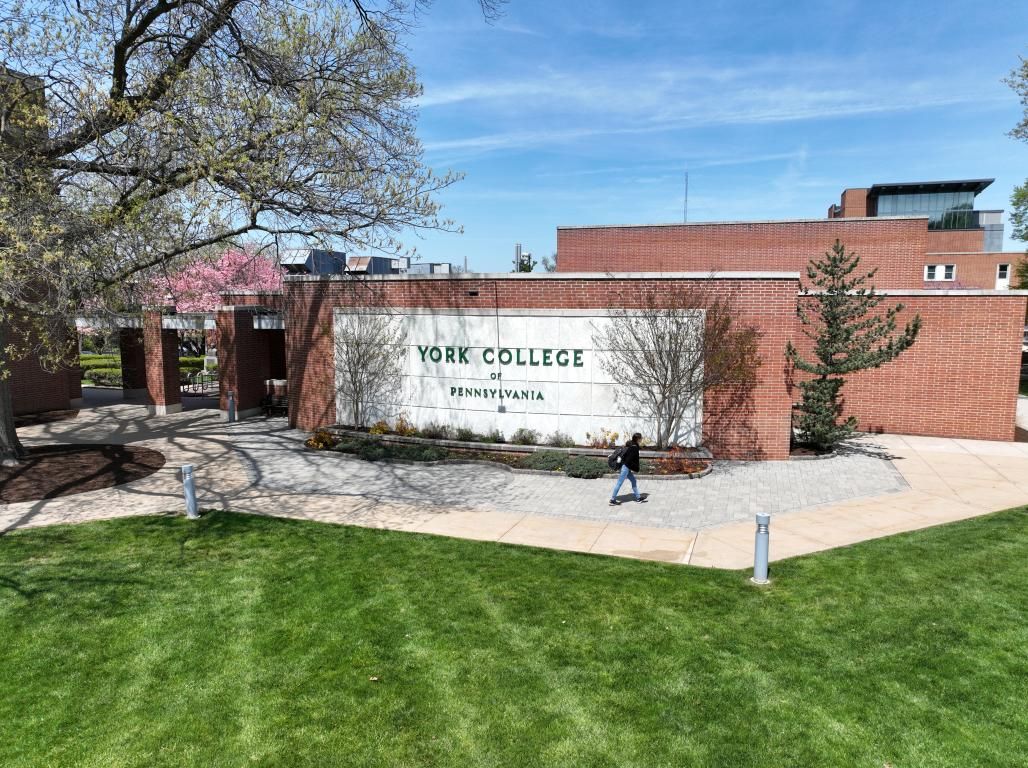 Exterior signage for York College of Pennsylvania displayed on a brick building on a sunny day. A green lawn and blue sky are visible, as well as a person walking on a sidewalk in front of the building.