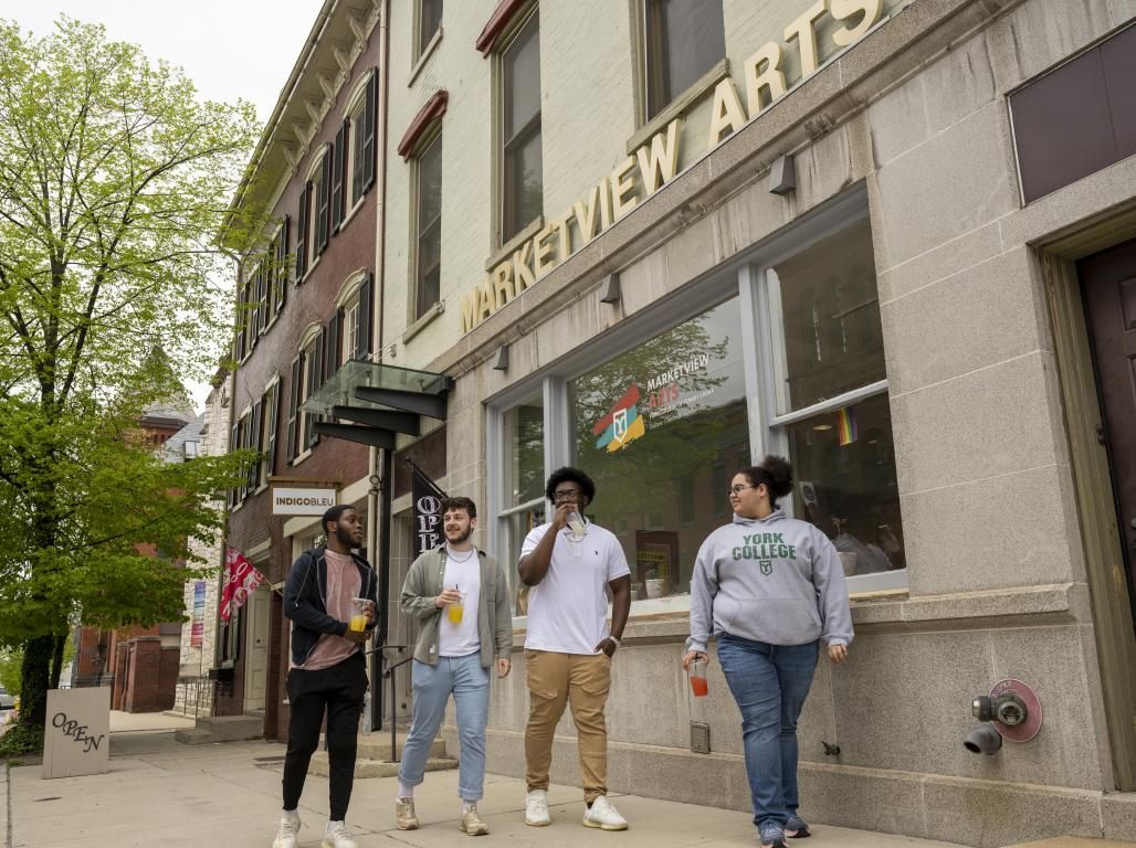 Four people walking on a city sidewalk, each holding drinks. A building with a sign reading "Marketview Arts" is visiible beside them.