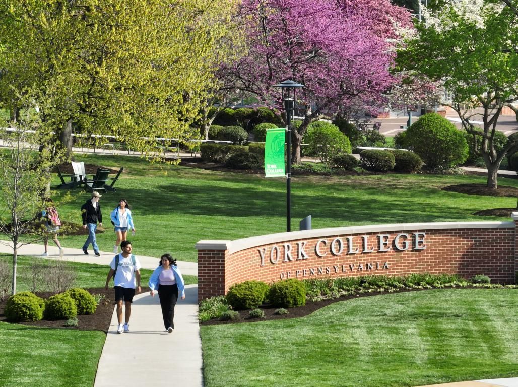 Students walking on a sidewalk on a sunny day at York College of Pennsylvania. Spring trees and college signage are visible.