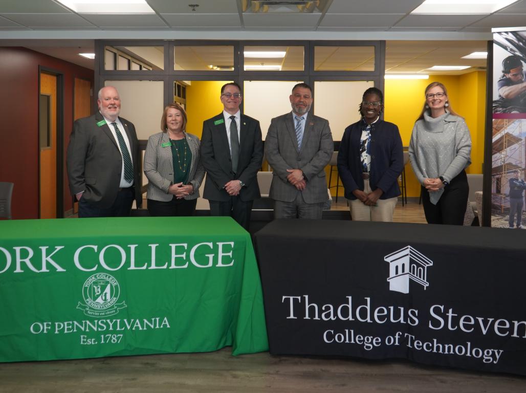 A group of people pose for a photo behind two tables with tablecloths showing logos for York College and Thaddeus Stevens.