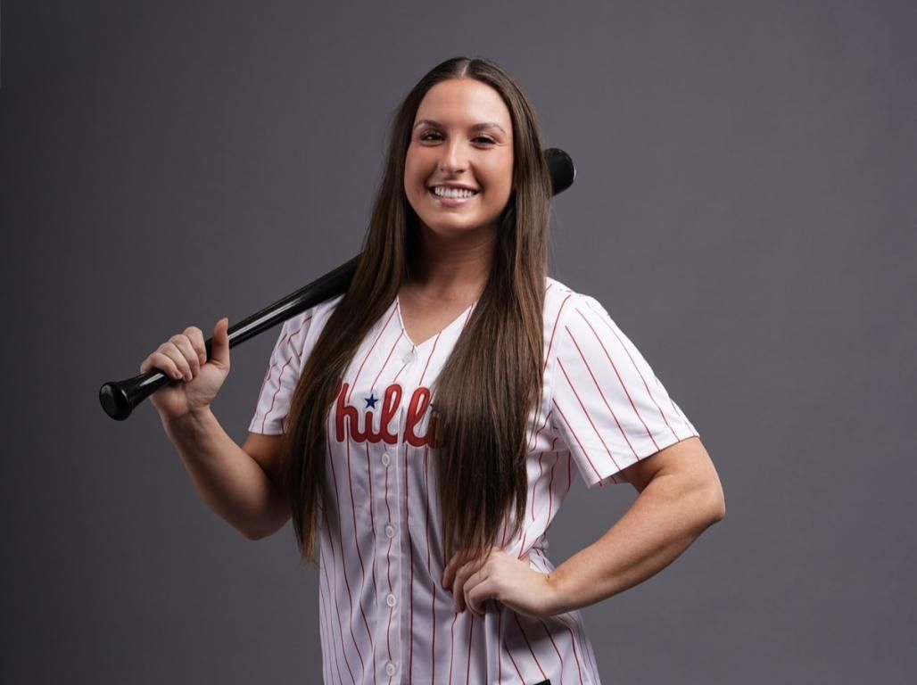 Person wearing a Phillies jersey smiles at the camera with a baseball bat balanced on their shoulder.