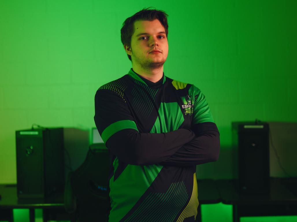 Person wearing a green and black ESports shirt poses with arms crossed in a dimly lit green room with computers visible in the background.