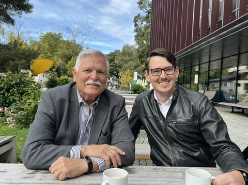Professor Charles (Chip) Kauffman poses with a student at a European university. They are seated at an outdoor table with coffee mugs.