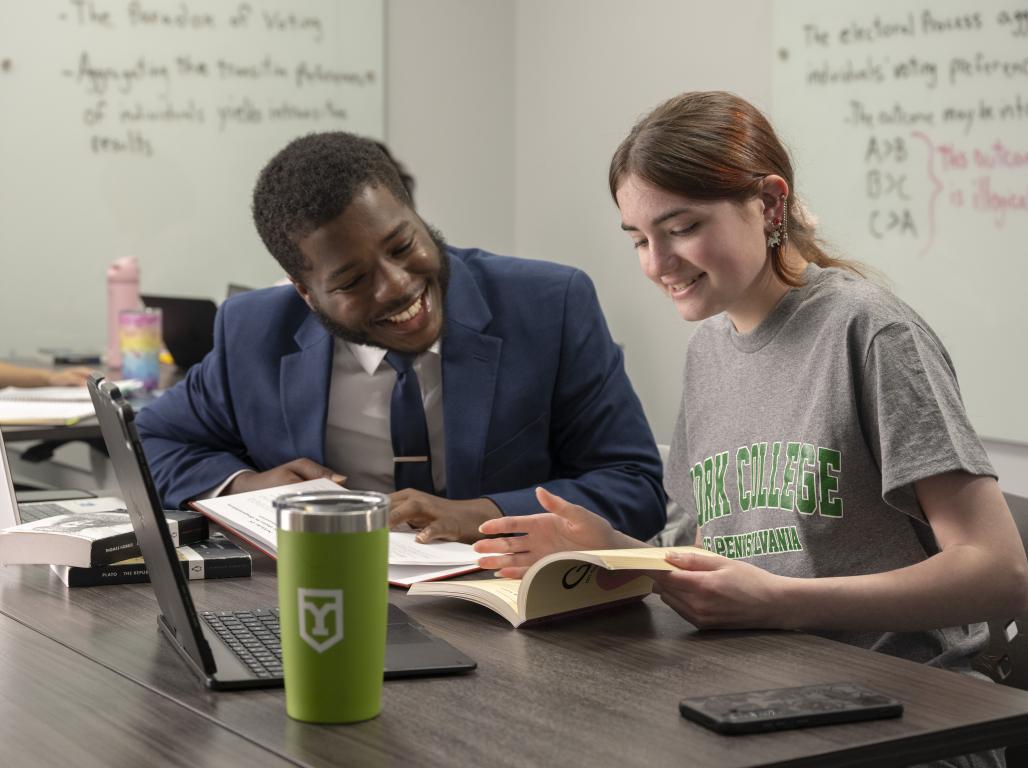 Two students look at a book together in a classroom with white boards behind them.