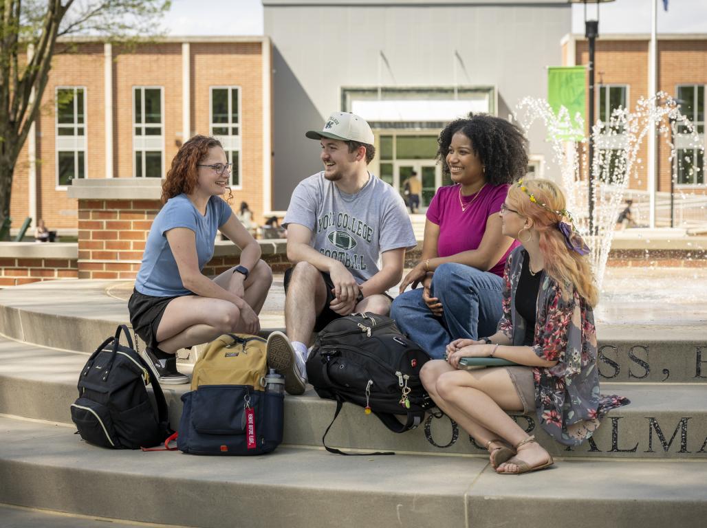 Four students sit next to the fountain talking with bookbags in front of them and the library behind them in the distance.