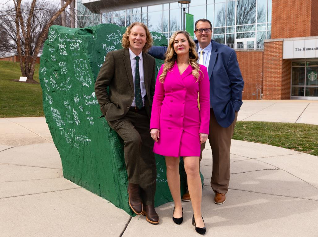 Three people stand posed together in professional clothes in front of a large green rock.