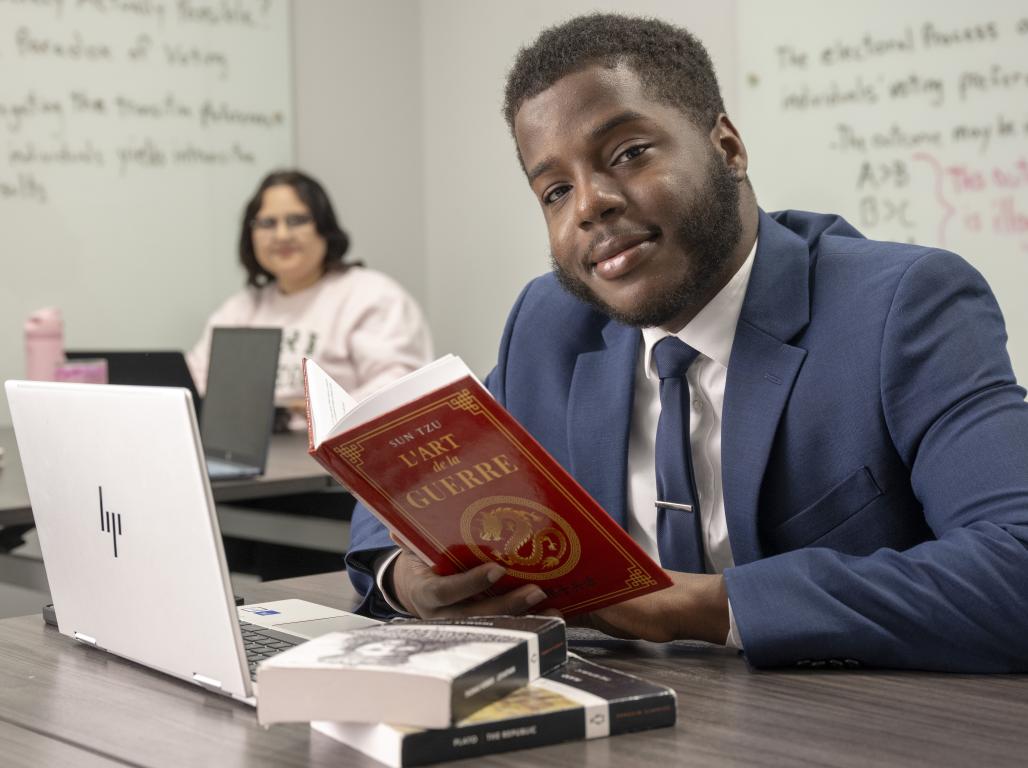 A student in a suit sits at a classroom table holding a book, with a laptop and additional books in front of him and another student seated in the background.